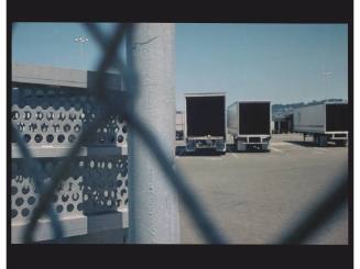 A color photograph taken through a chain-link fence of parked semi-trucks.

