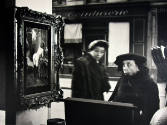 A black and white photograph of two women peering into a store window containing a painting of …