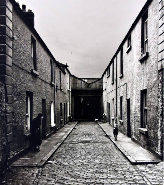 A black and white photograph of a cobblestone street flanked by buildings with an older woman i…