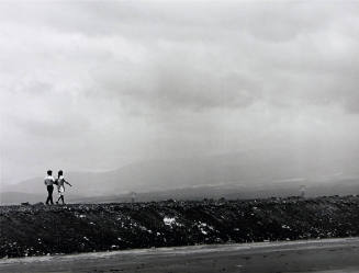 A black and white photograph of a couple walking along a road with mountains in the background.