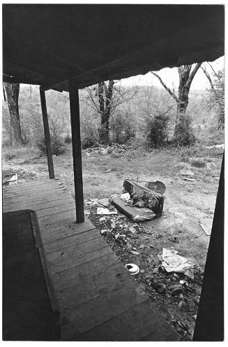 A black and white photograph taken from the porch of a home with a dog laying on a dilapidated …