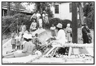 A black and white photograph of women seated on top of a car decorated with floral garlands in …