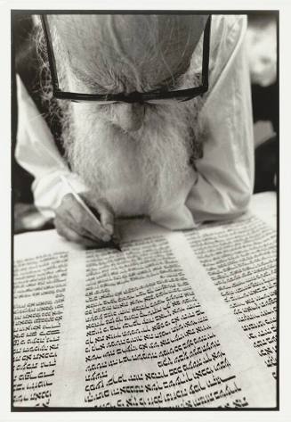 A man writing on a document with three columns of text written in Hebrew.