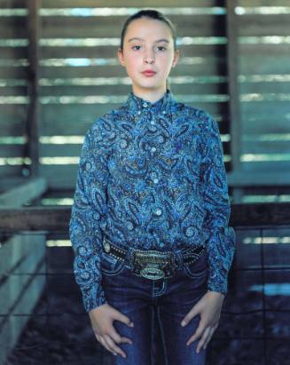 A young girl stands in a barn with her hands by her side dressed in a blue paisley shirt and bl…