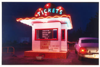 A color photograph of a drive-in theater ticket booth illuminated by a plethora of neon lights.…