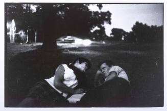 A black and white photograph of a man and woman lying in the foreground of a park at dusk, punc…