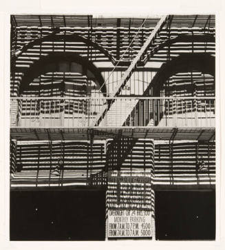 A black and white photograph shows a heavily-shadowed building with a fire escape in the center…