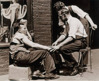 A black and white photograph of three men conversing on a sidewalk in front of a store window w…