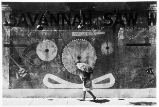 A black and white photograph of a woman walking beside the old Savannah Saw Works building. 