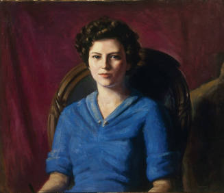 Portrait of a young woman, wearing a blue blouse and sitting in a green upholstered chair. The …