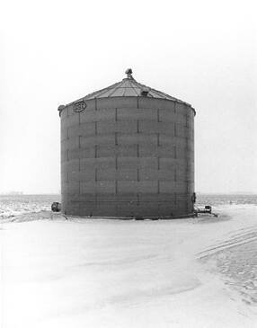 A black and white photograph of a grain silo or storage bin in a field.

