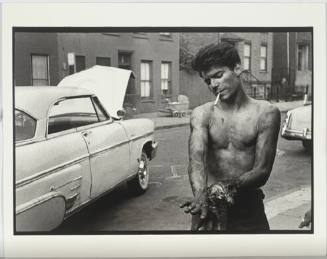 A black and white photograph of a young man wiping oil off his hands by a car with the hood rai…