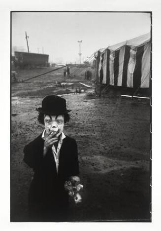 A black and white photograph of a little person wearing clown make-up and smoking outside of a …