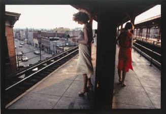 A color photograph of two women standing and reading in brightly colored dresses while waiting …