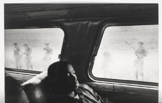 A black and white photograph of a young man seated on a bus with American soldiers visible thro…