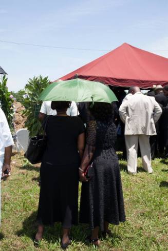 Two women in long black dresses stand together underneath a green umbrella in a cemetery in fro…