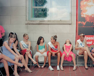 Girls in pageantry dresses, some with tiaras and sashes, sitting in a row on folding chairs. 