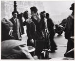 A black and white photograph of a couple kissing on a busy sidewalk by a cafe.