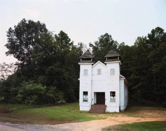 A white wooden church on a sandy and grassy plot surrounded by trees.

