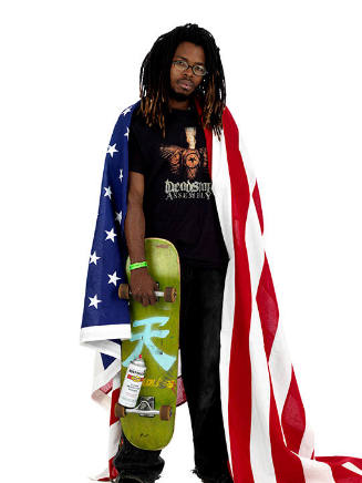 A young man with dreadlocks and glasses holding a skateboard in his right hand with the America…