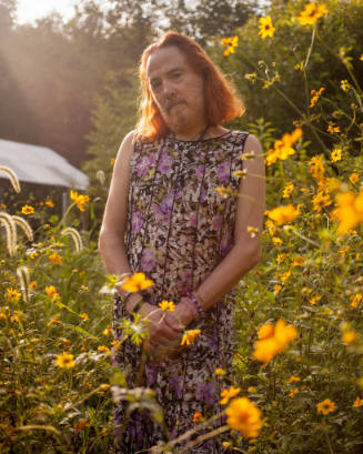 A man with a beard and long red hair is standing in a field of yellow wildflowers wearing a pur…