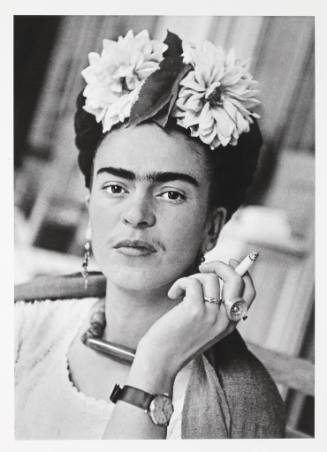 Frida Kahlo looks directly at the camera with a cigarette in her left hand and a large flowered…