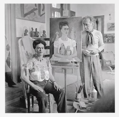 Frida is sitting in a chair in front of her painting “Me and my Parrots” looking towards the ca…