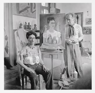 Frida is sitting in a chair in front of her painting “Me and my Parrots” looking towards the ca…