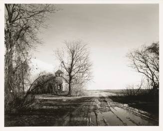 A muddy dirt road leading to a decrepit church located on the left-hand side in a landscape wit…