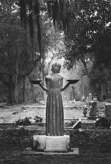 A black and white photograph of a statue in a cemetery. The statue is of a young girl on a plin…