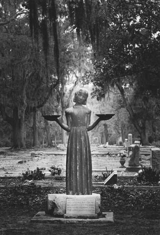 A black and white photograph of a statue in a cemetery. The statue is of a young girl on a plin…