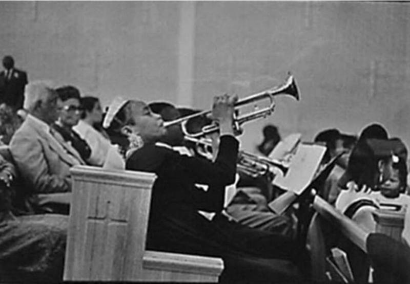 A black and white photograph of a young person sitting in a church pew playing a trumpet.