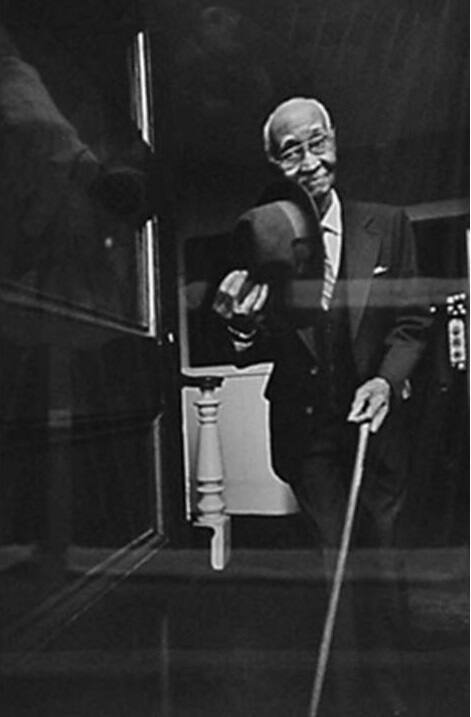 A black and white photograph of a man in a dark suit, holding a fedora in one hand and a cane i…