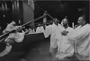 A black and white photograph of a man being baptized with microphones hovering over the baptism…