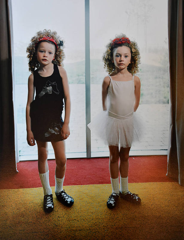 A color photograph of two blonde girls in ballet garments standing by a curtained window.