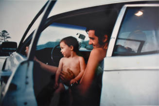 A color photograph of a man with a child in his lap, seated in the driver's seat of a car with …