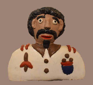 A painted cement sculpture of a bust of a man with black curly hair, mustache and beard wearing…