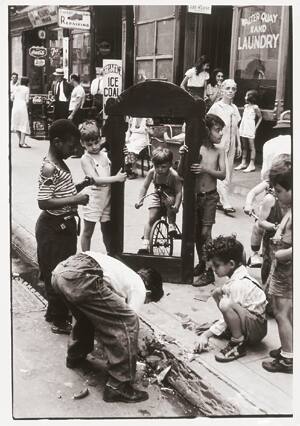A black and white photograph of children playing with a broken mirror frame and a tricycle on a…