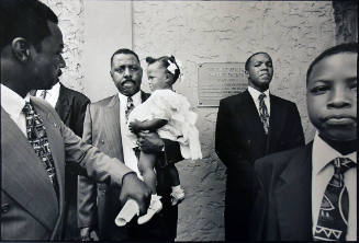 A black and white photograph of a man holding a little girl in a white dress with a young boy p…