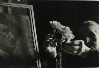 A woman emerges from the background to touch a white flower in a vase next to a portrait painti…