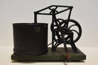 A cast iron flywheel food processor attached to a wooden stand with a tin bowl, gears and woode…