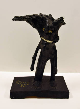 An anthropomorphic sculpture in solid black with three limbs, a string of white beaded teeth an…