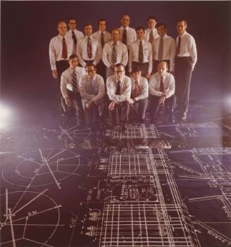 A group of men in white shirts with black ties standing on blueprints.