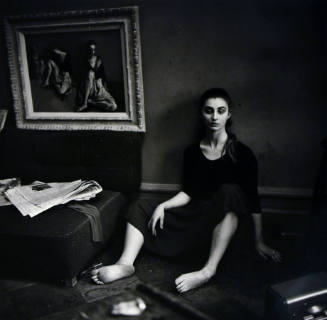 A black and white photograph of a woman sitting on the floor with a low hung painting beside he…