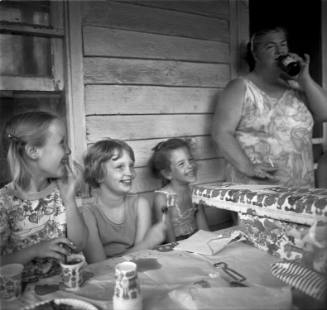 A black and white photograph of three children and one woman on the back porch of a wood clad h…