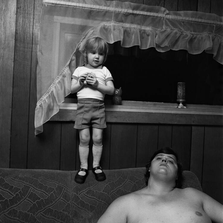 A black and white photograph of a child standing on the back of a sofa where a man is seated.