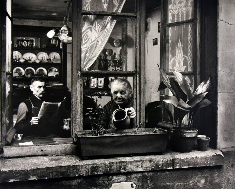 A black and white photograph of a woman watering the flowers in the box outside her window. Ins…