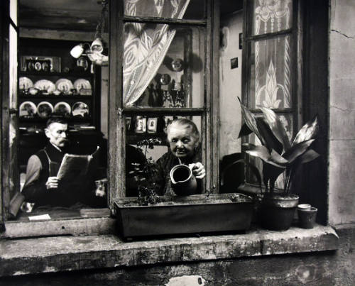 A black and white photograph of a woman watering the flowers in the box outside her window. Ins…