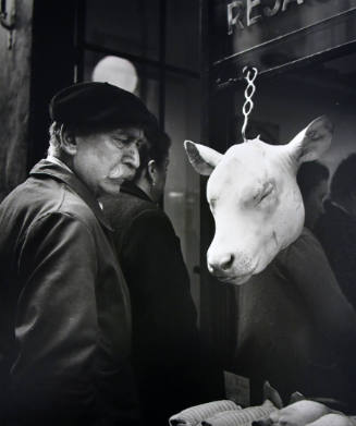 A black and white photograph of a man in a beret looking at a white cow's head on a hook.