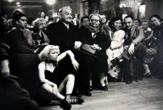 A black and white photograph of seated individuals with a blonde woman sitting on the floor at …